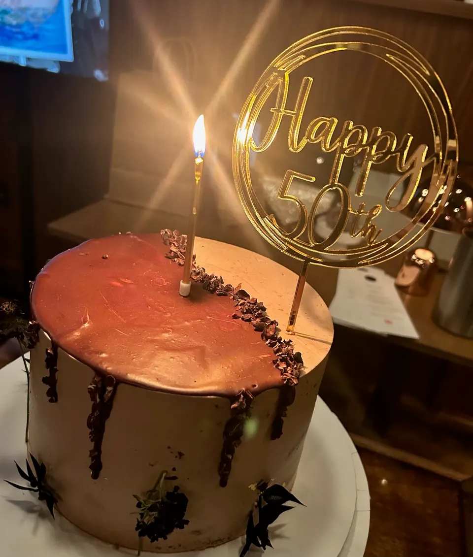 A tall, round chocolate drip cake with a single lit candle and a gold 'Happy 50th' topper, set on a white plate.