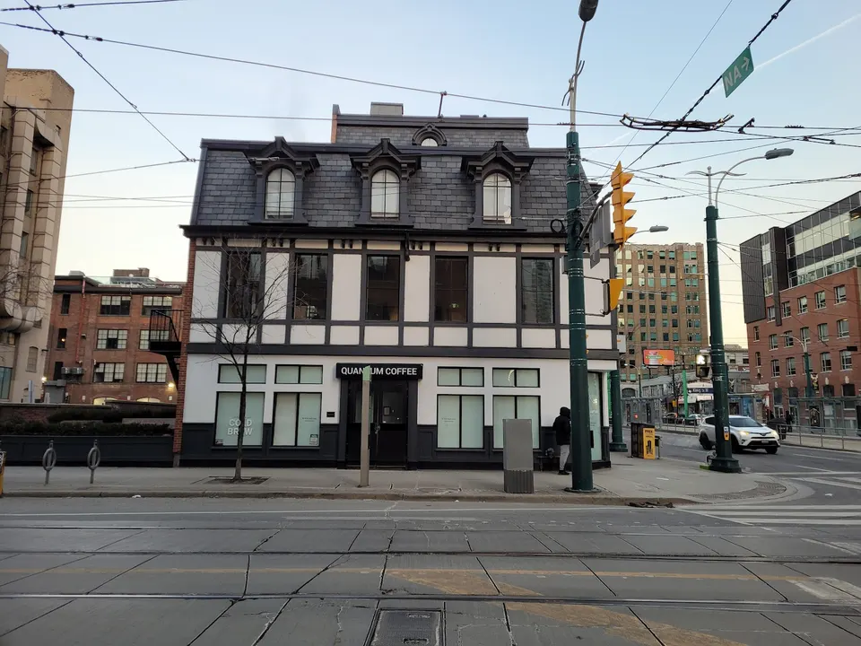 A two-story building with a dark gray mansard roof and white and dark gray facade, housing Quantum Coffee. The street in front has tram tracks and a crosswalk. Utility poles and wires crisscross the sky. Other buildings are visible in the background.