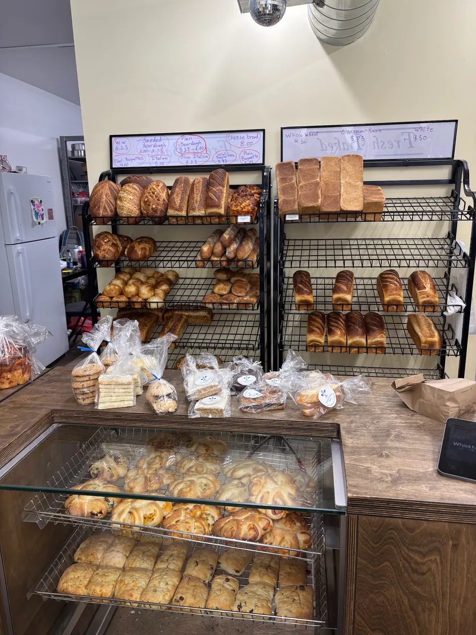 A bakery display featuring wire racks filled with freshly baked sourdough, baguettes, and sandwich loaves, with packaged cookies on the counter and pastries in a glass display case below.