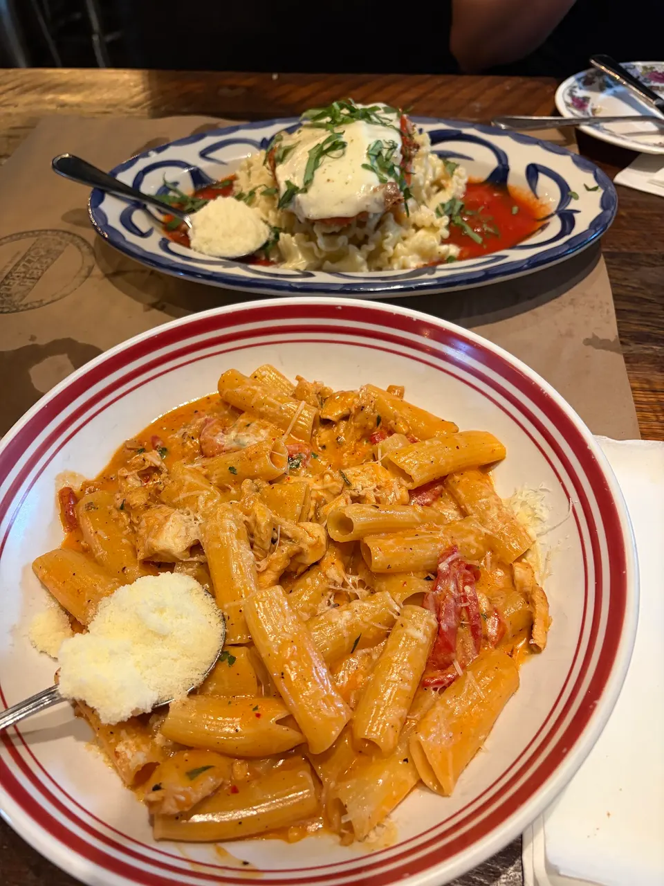 A close-up of a bowl of rigatoni pasta in a creamy tomato sauce with a spoon of grated cheese, with another pasta dish in a blue and white plate in the background.