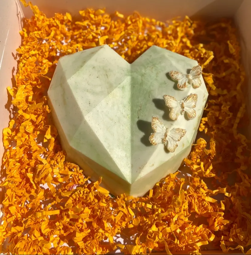 A geometric, heart-shaped green and white marbled chocolate cake decorated with three small white edible butterflies, resting in a box filled with shredded orange paper.