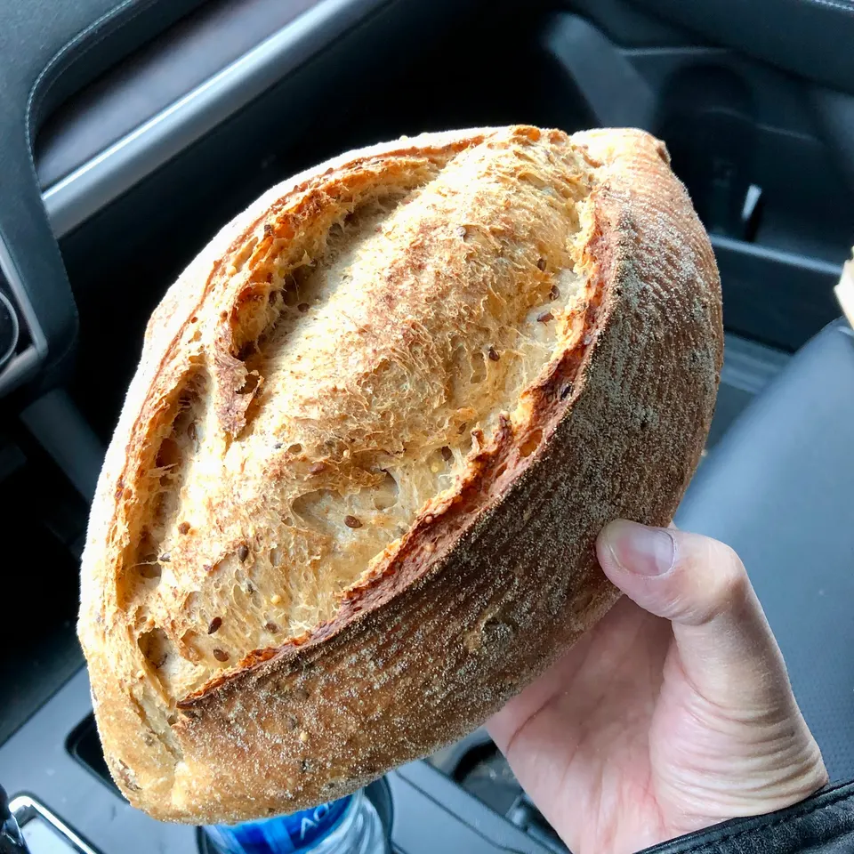 A hand holding a large, freshly baked loaf of sourdough bread with a golden-brown, textured crust inside a car.