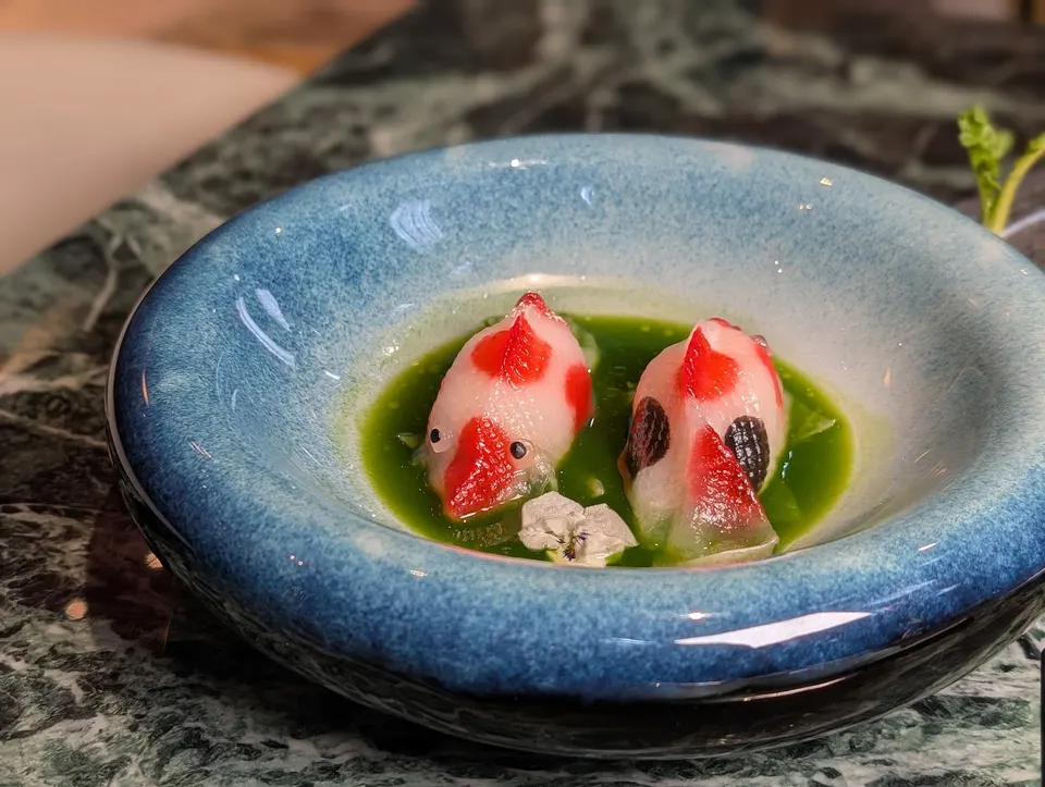 Two delicate, fish-shaped dim sum dumplings resembling koi fish, decorated with red and black spots, served in a shallow pool of vibrant green broth inside a blue ceramic bowl.