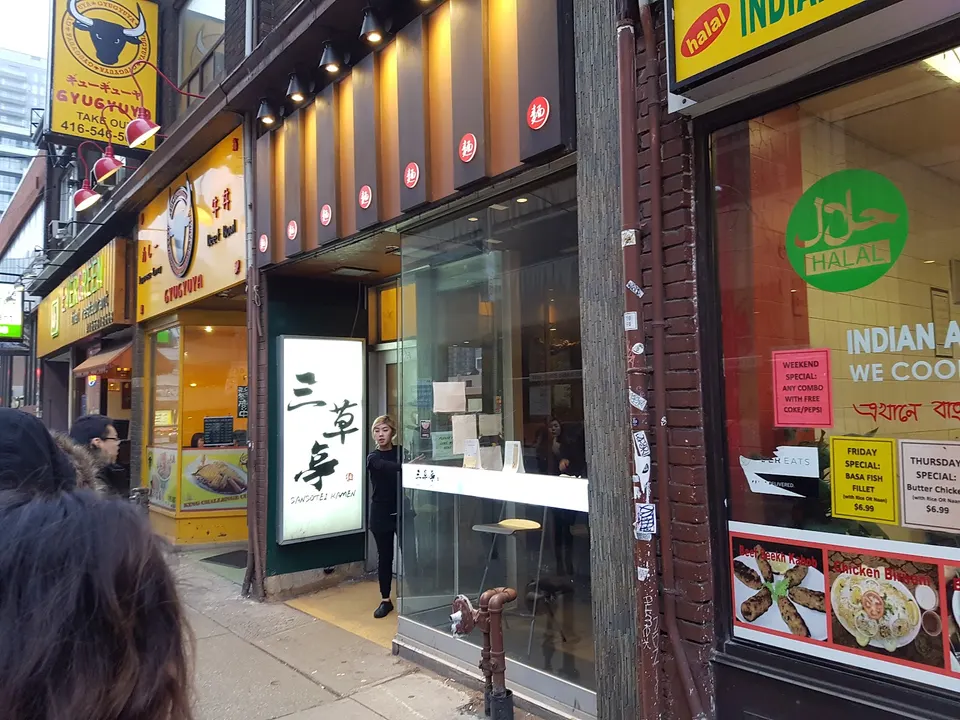 Street view of the Sansotei Ramen storefront with its distinctive white vertical sign featuring Japanese calligraphy, situated between other colorful restaurants on a city sidewalk.