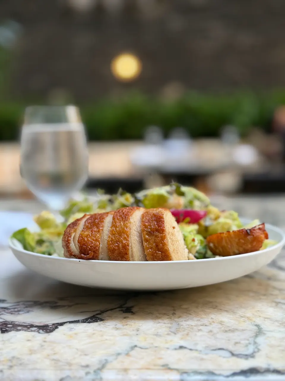 A sliced roasted chicken breast served over a fresh green salad in a white bowl, set on a marble table with a water glass in the blurred background.