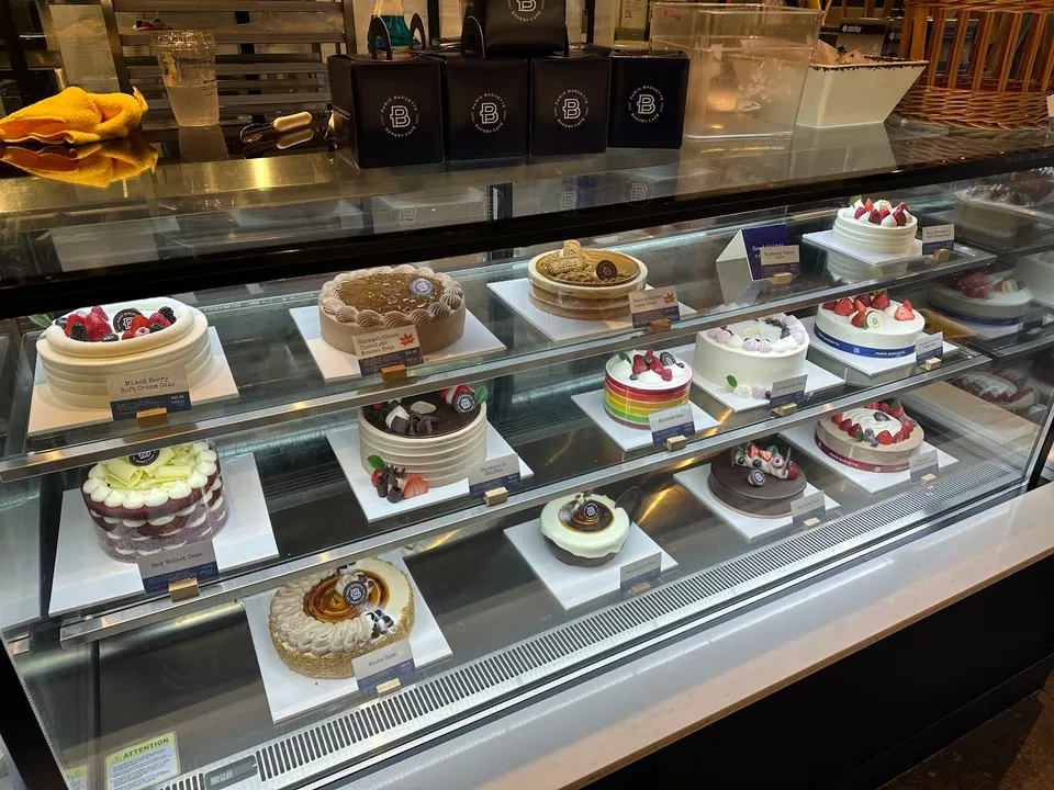 A glass display case filled with various decorated whole cakes on three shelves at Paris Baguette, including fruit-topped, chocolate, and cream cakes.