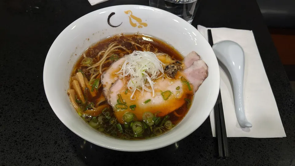 A bowl of shoyu ramen with sliced chashu pork, green onions, bamboo shoots, and shredded white leeks, served with chopsticks and a spoon on a black table.