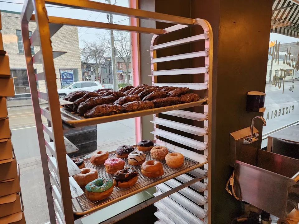 A metal baker's rack inside a pizzeria window displays a tray of dark chocolate pastries on top and a wire rack of assorted glazed and frosted donuts below.
