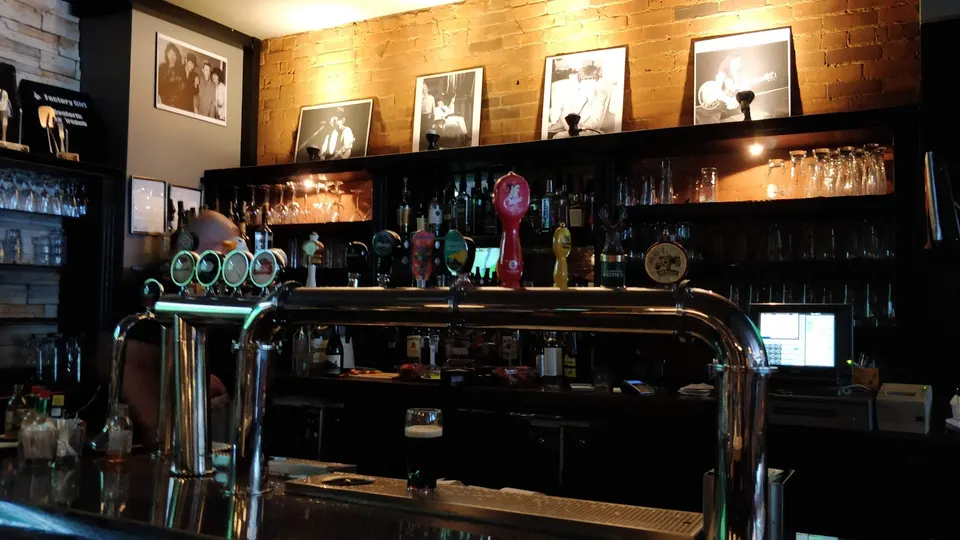 A close-up view of a dark wood bar counter with shiny chrome beer taps, a single dark pint of beer, and shelves of liquor bottles and glassware under warm lighting.