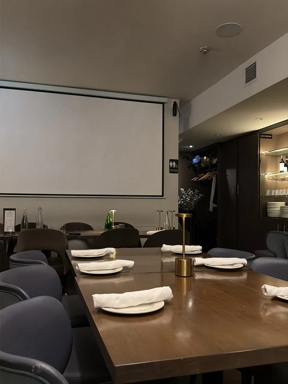 A long wooden dining table set with white plates, rolled napkins, and gold table lamps, surrounded by grey upholstered chairs in a dimly lit restaurant dining room with a large projection screen on the wall.