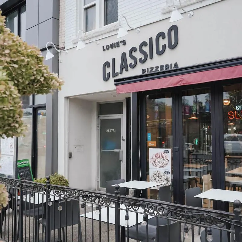 The storefront of Louie's Classico Pizzeria featuring a white facade, black signage, a red awning, and an outdoor patio area with tables and chairs behind a black metal fence.