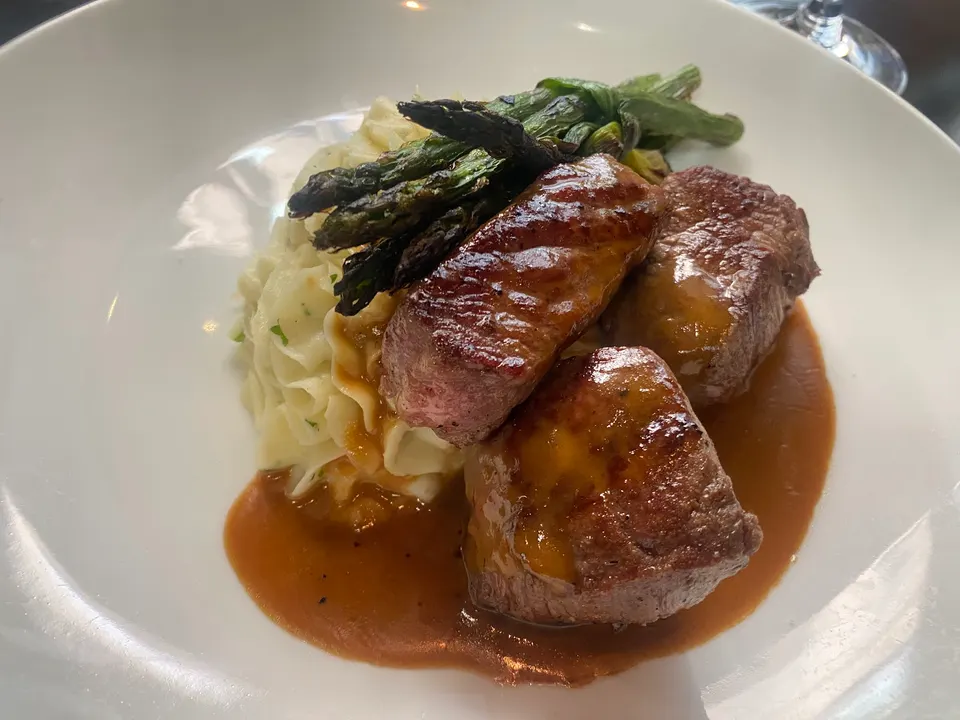 Three thick cuts of seared steak served over mashed potatoes with grilled asparagus and a rich brown sauce in a white bowl.