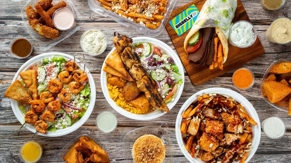 A flat lay of Greek street food dishes on a wooden table, including a gyro wrap, chicken souvlaki bowl, shrimp salad, loaded fries, pita bread, and various dipping sauces.