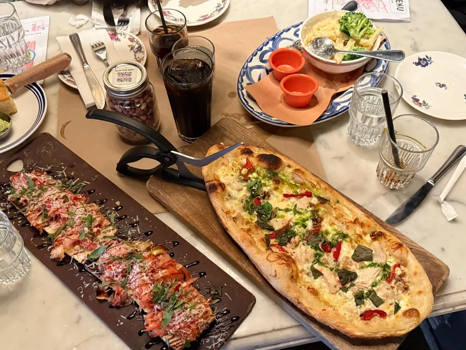 A table spread at Scaddabush Italian Kitchen & Bar Scarborough featuring two long flatbread pizzas on wooden boards, a bowl of pasta with broccoli, dipping sauces, and cold drinks.