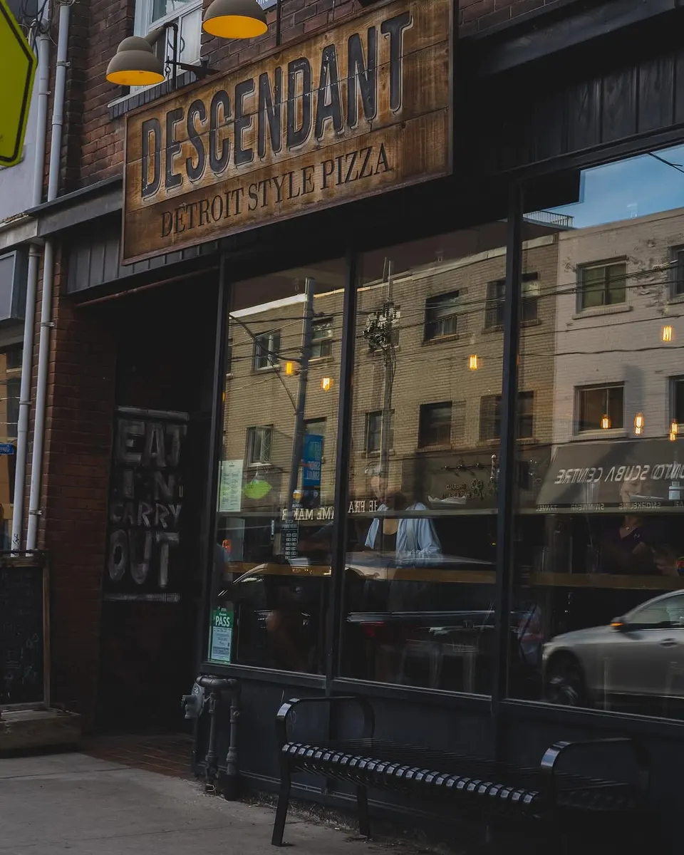 The exterior storefront of Descendant Detroit Style Pizza, featuring a rustic wooden sign, large glass windows reflecting the street, and a black metal bench on the sidewalk.