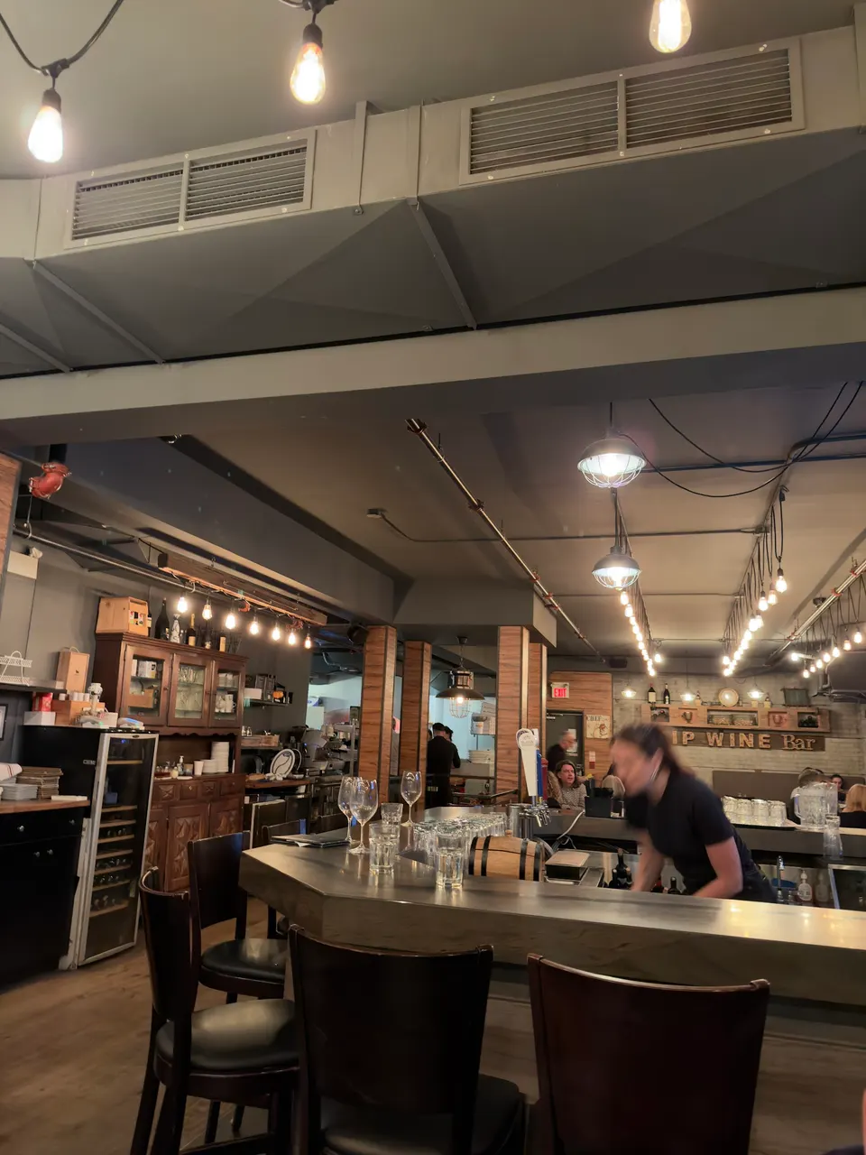 Interior view of SIP Wine Bar showing a modern curved bar counter with wooden bar stools, hanging pendant lights, and a bartender working behind the counter.