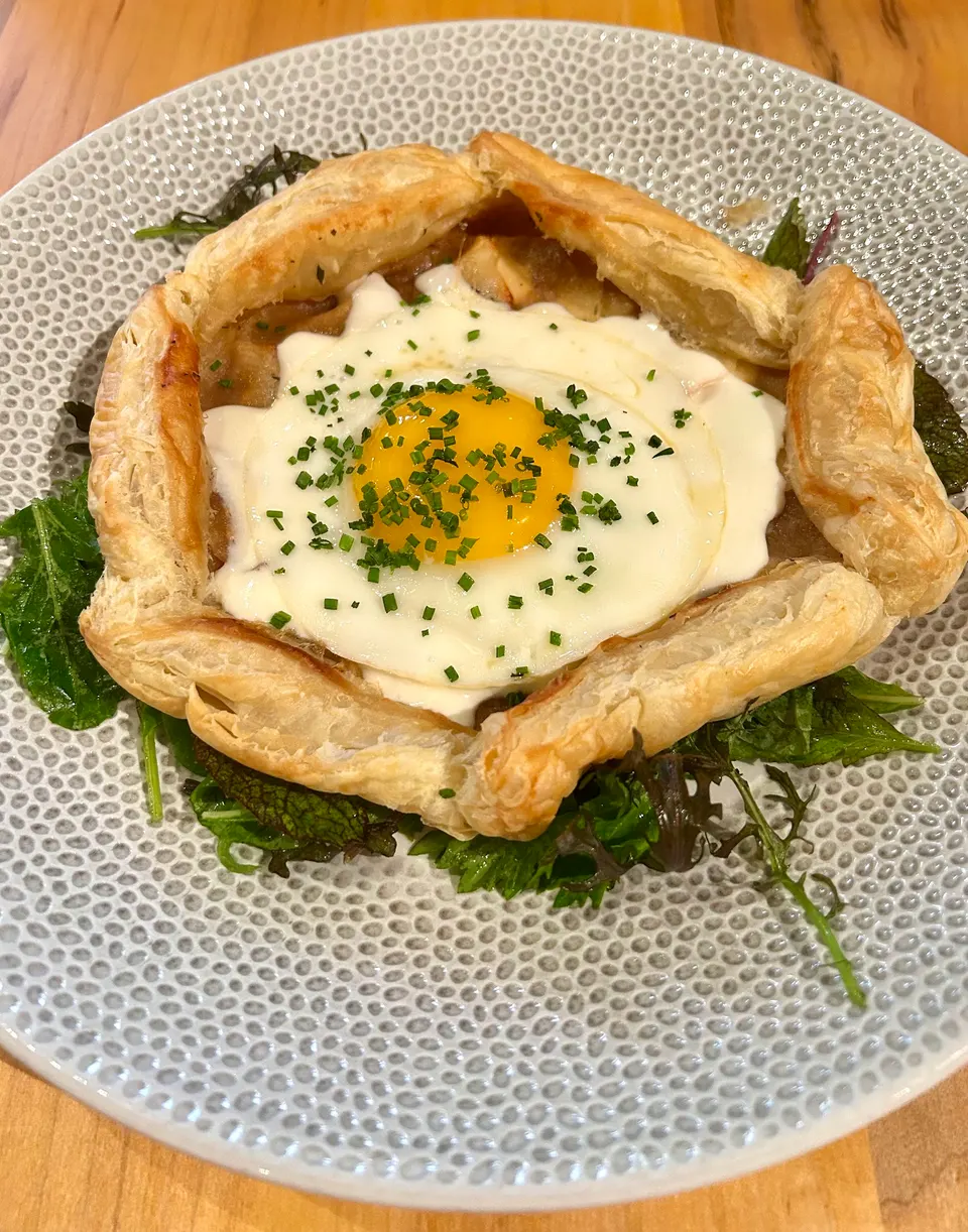 A savory puff pastry tart filled with a fried egg topped with chopped chives, served on a bed of mixed greens on a textured grey plate.