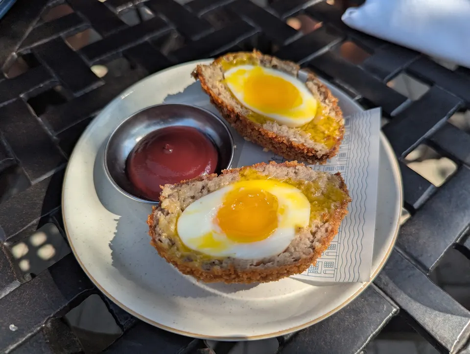 Two Scotch eggs, cut in half to reveal soft-boiled yolks, served on a white plate with a small metal cup of dark red sauce, all resting on a black metal outdoor table.