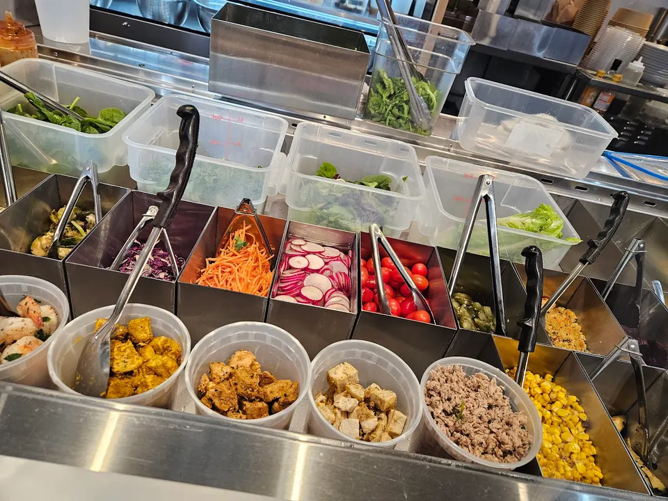 A salad bar counter at Green Box featuring various fresh ingredients in metal and plastic containers, including spinach, carrots, radishes, cherry tomatoes, corn, and proteins like chicken and tofu.