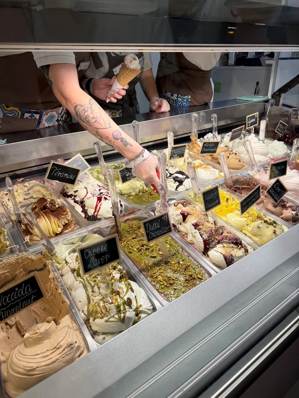 A colorful display case filled with various gelato flavors like Hazelnut, Cheese Lover, Dubai Chocolate, and Tiramisu, with a server holding a cone in the background.
