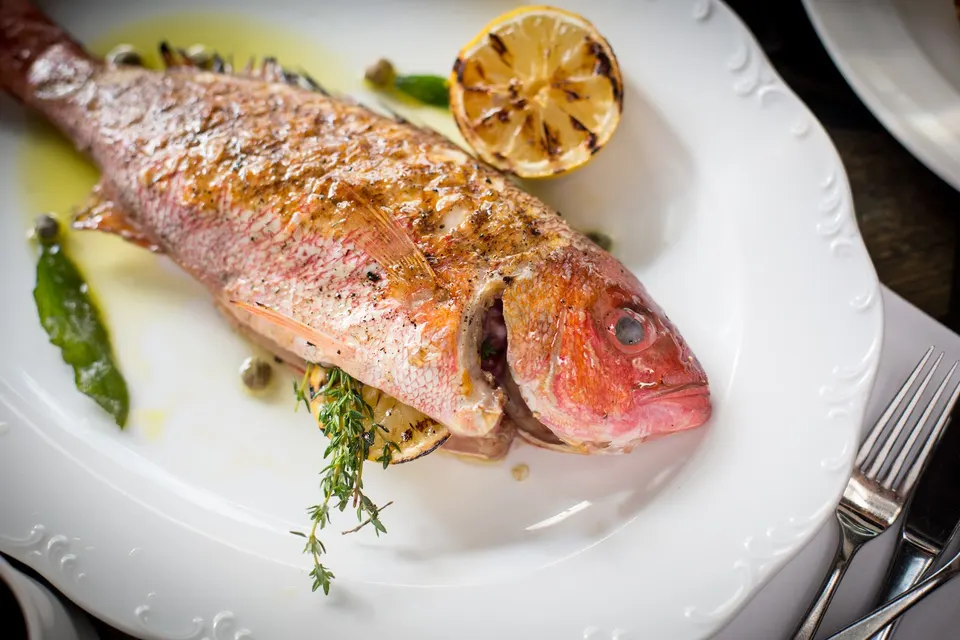 A whole grilled red snapper served on a white platter, garnished with a grilled lemon half, fresh herbs, capers, and olive oil.
