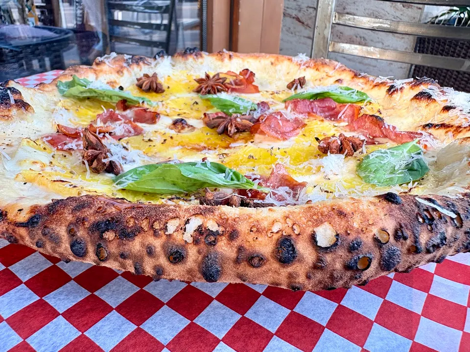 A wood-fired pizza with a charred, bubbly crust topped with yellow sauce, prosciutto, fresh basil leaves, grated cheese, and whole star anise, served on a red and white checkered tablecloth.