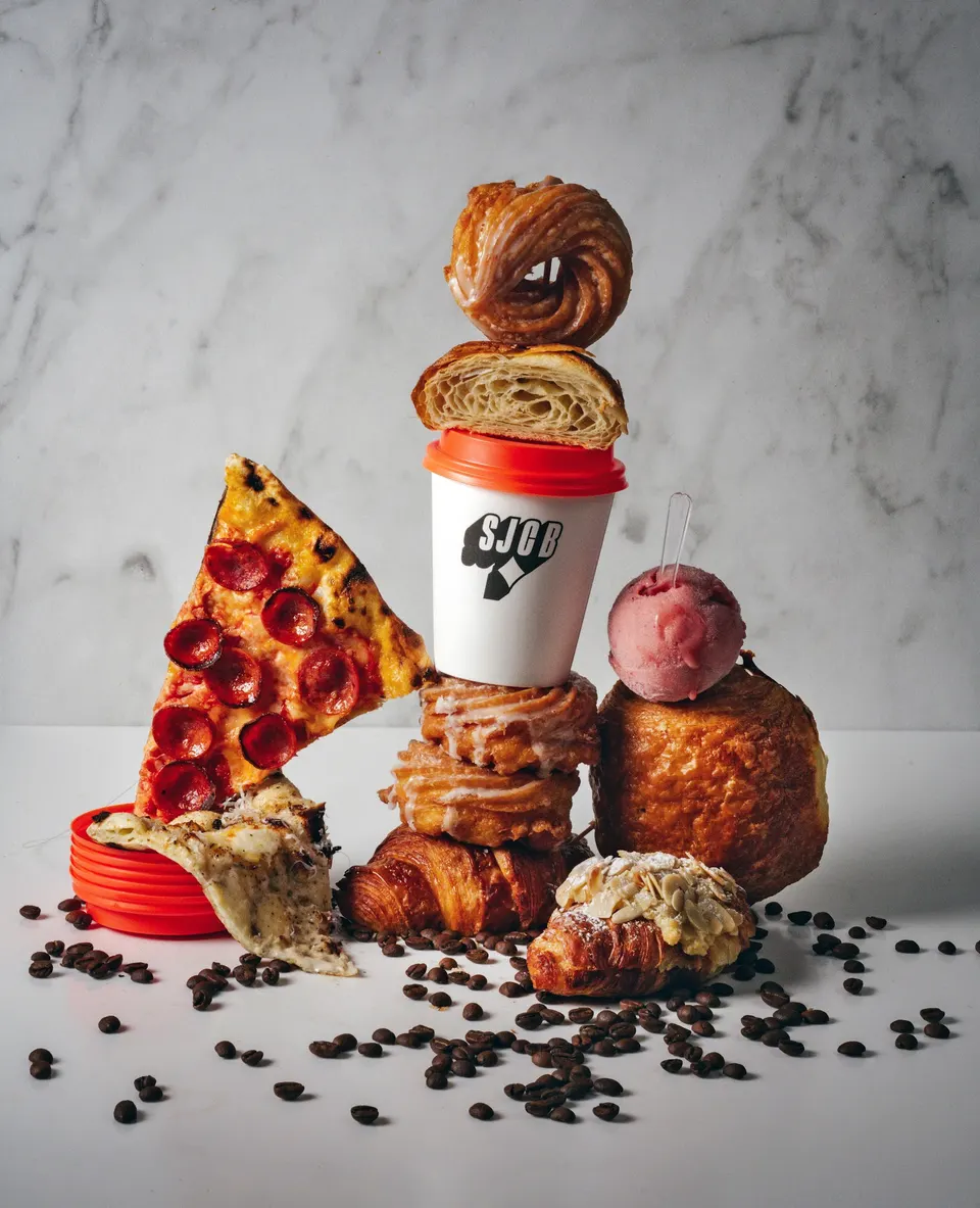 A creative stack of food items featuring a slice of pepperoni pizza, a coffee cup, croissants, pastries, and a scoop of pink gelato, surrounded by coffee beans on a white surface.