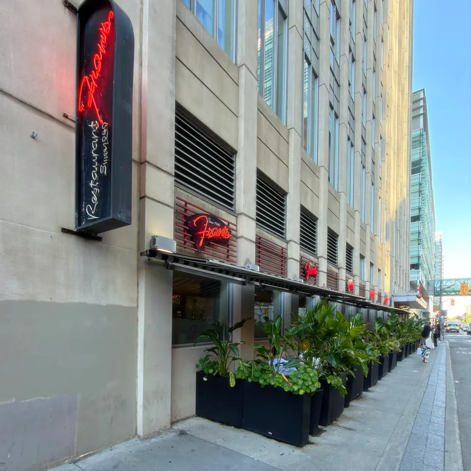 Exterior view of Fran's Restaurant featuring a sidewalk patio lined with lush green planters, a black awning, and glowing red neon signs on a modern city building facade.