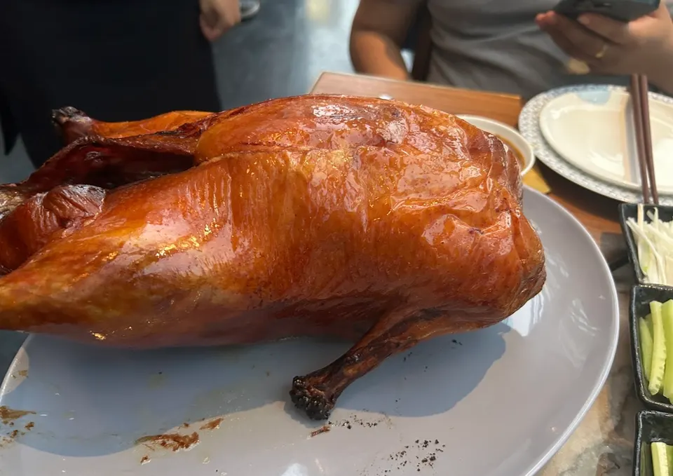 A whole, golden-brown roasted Peking duck with crispy skin served on a large grey platter at a dining table with side dishes.