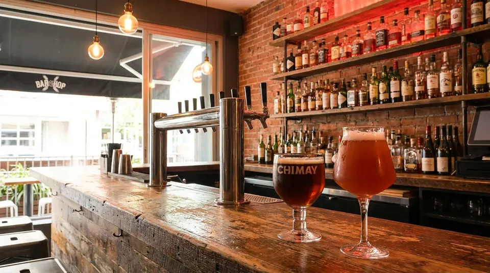 Two glasses of craft beer, one dark and one amber, sit on a rustic wooden bar counter in front of draft beer taps and a brick wall lined with liquor bottles.