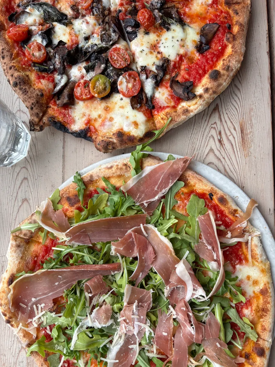 Top-down view of two wood-fired pizzas on a rustic wooden table. The top pizza features mushrooms, cherry tomatoes, and melted mozzarella. The bottom pizza is topped with fresh arugula, prosciutto, and shaved parmesan.