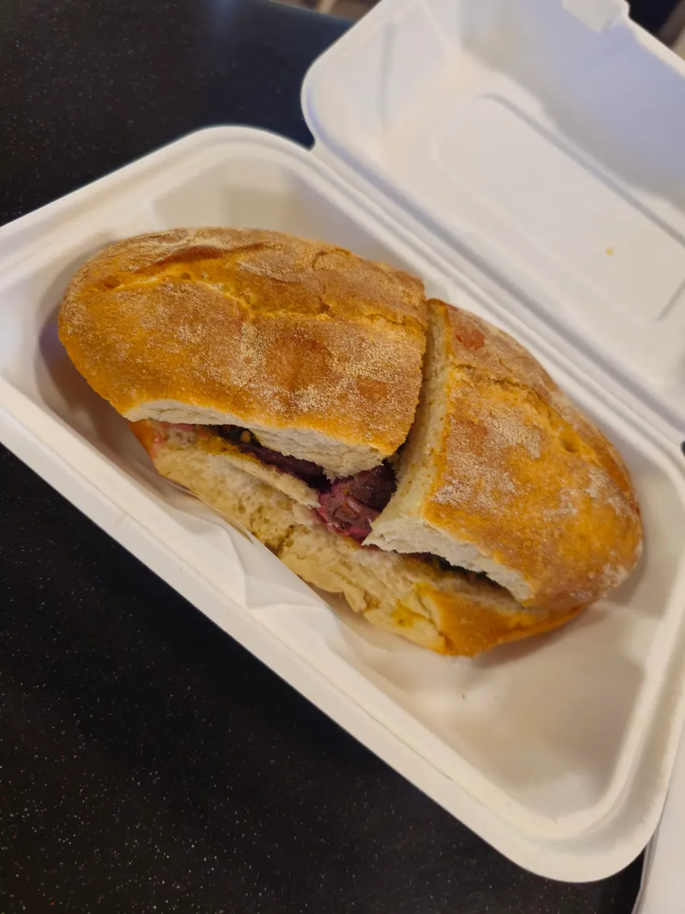 A hearty sandwich with crusty bread and meat filling, cut in half and placed in a white takeout container on a dark surface.
