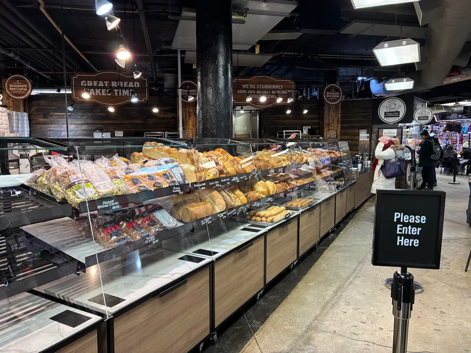 A long bakery display counter at Stonemill Bakehouse Ltd filled with various loaves of bread, buns, and packaged baked goods under warm lighting, with a 'Please Enter Here' sign in the foreground.