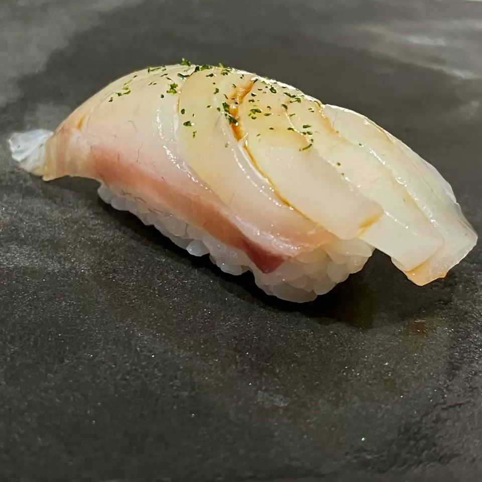 A single piece of nigiri sushi topped with translucent white fish, a light glaze, and a sprinkle of green herbs, served on a dark textured slate plate.