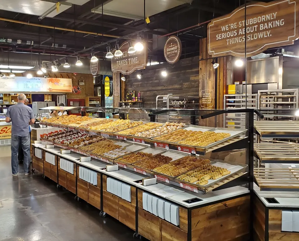 A large bakery display case filled with rows of pastries, croissants, and baked goods under warm lighting, with wooden signs reading 'We're stubbornly serious about baking things slowly' and 'Better bread takes time.'