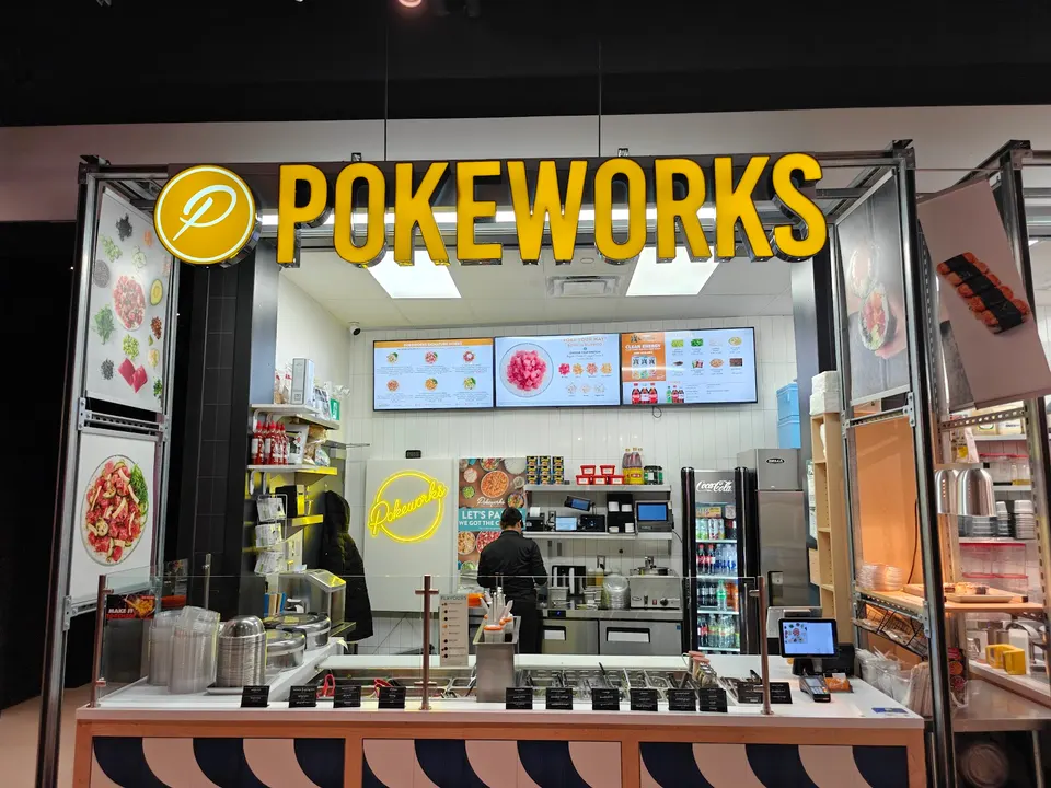 The front counter and interior of a Pokeworks restaurant. A large yellow 'POKEWORKS' sign hangs above a service counter. Digital menu boards display food items. Two staff members are visible behind the counter, preparing food. A refrigerator stocked with drinks is on the right.
