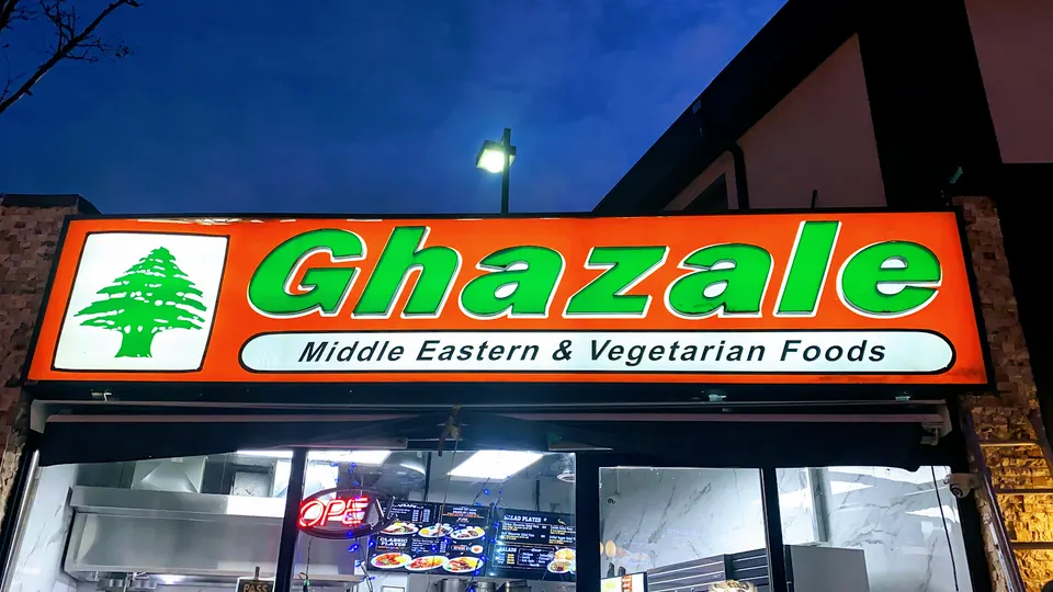 Bright orange and green illuminated storefront sign for Ghazale Middle Eastern & Vegetarian Foods at dusk.