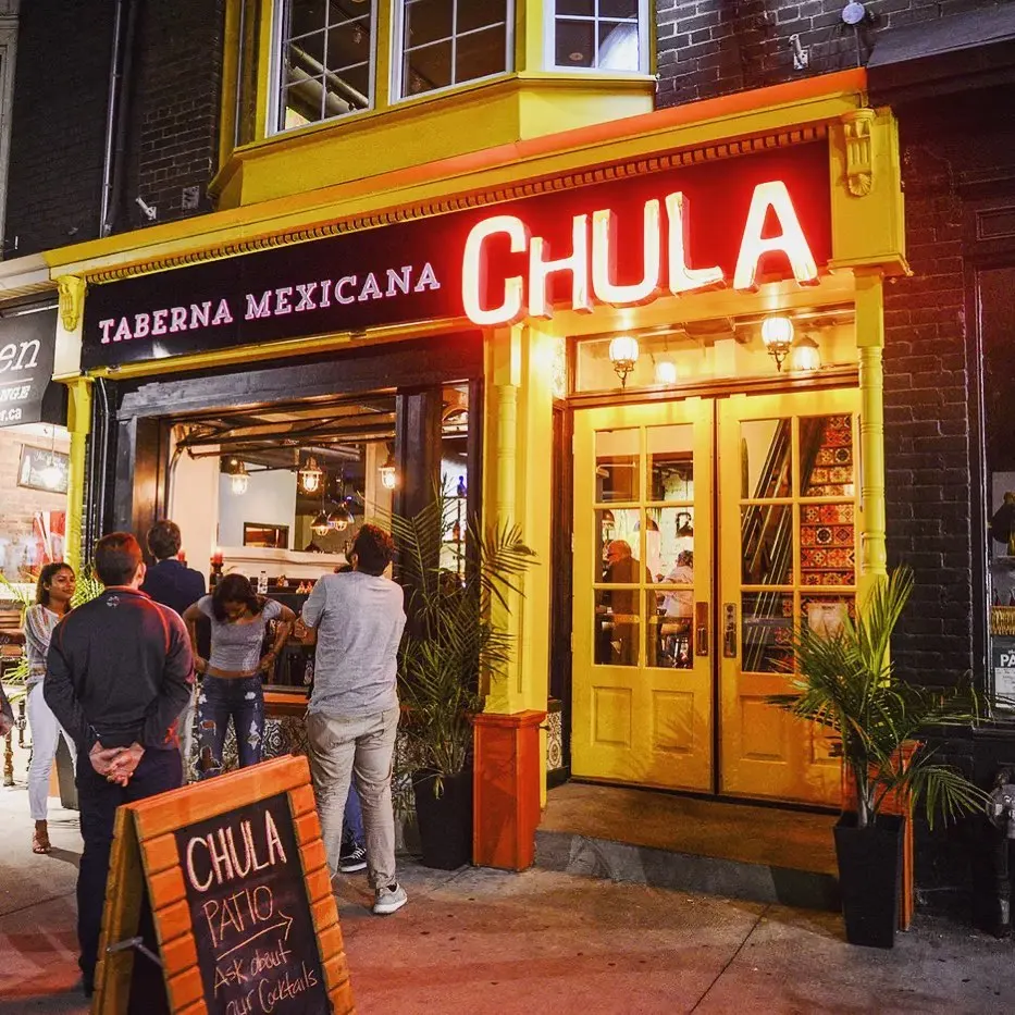 The vibrant yellow storefront of Chula Taberna Mexicana at night, featuring a glowing red neon sign, double glass doors, potted palm plants, and patrons gathered outside near a wooden sandwich board.