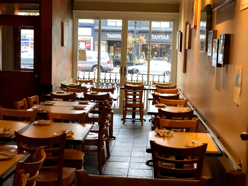 An empty dining room at Frank's Pizza House with wooden tables and chairs set with plates and cutlery, looking out through glass doors to a street view.