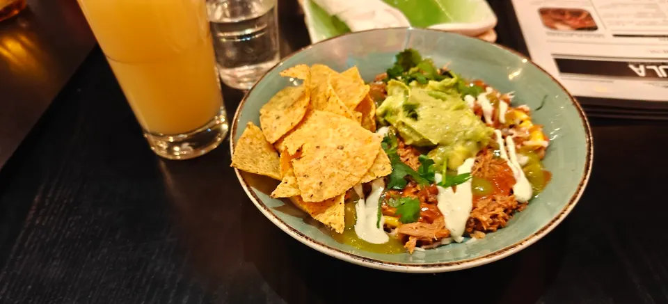 A bowl of Mexican pulled pork carnitas topped with guacamole, sour cream, and fresh cilantro, served with a side of tortilla chips next to a glass of juice.