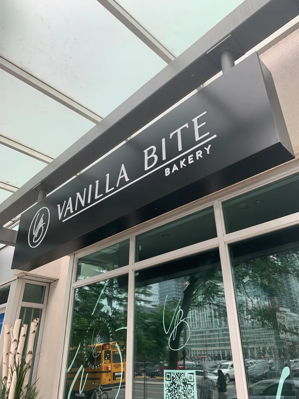 A black exterior sign with white lettering reads 'VANILLA BITE BAKERY' above a glass storefront reflecting city buildings and a yellow school bus.