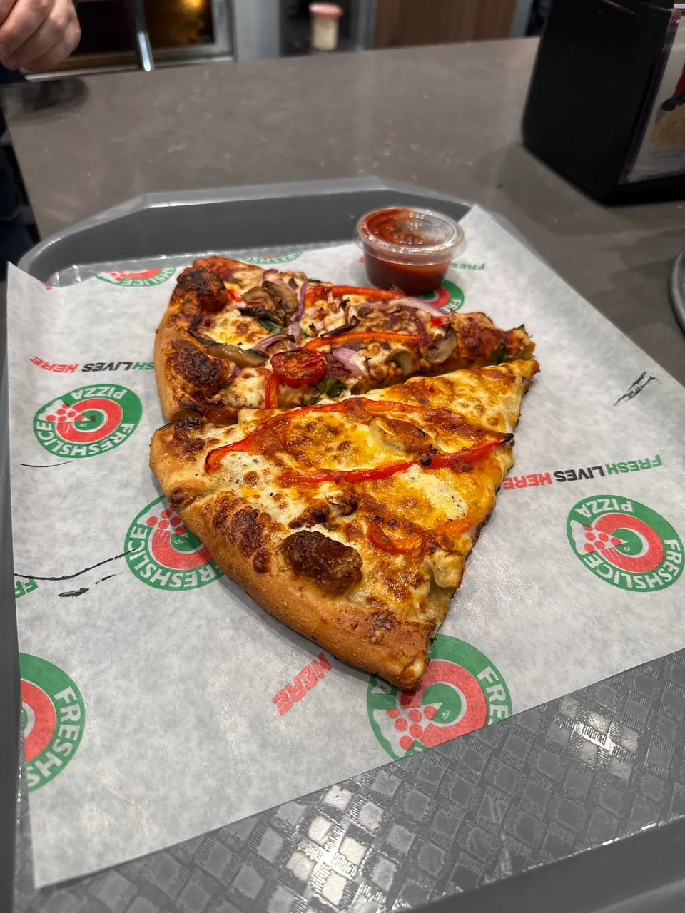 Two slices of pizza with toppings like red peppers, onions, and mushrooms served on branded paper on a tray next to a small cup of dipping sauce.