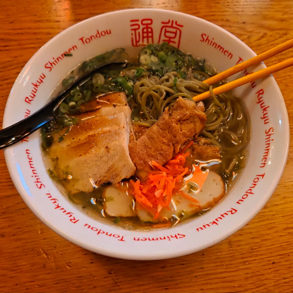 A bowl of ramen with green noodles, sliced pork belly, braised pork, pickled ginger, and fish cakes in a clear broth, served in a white bowl with red lettering.