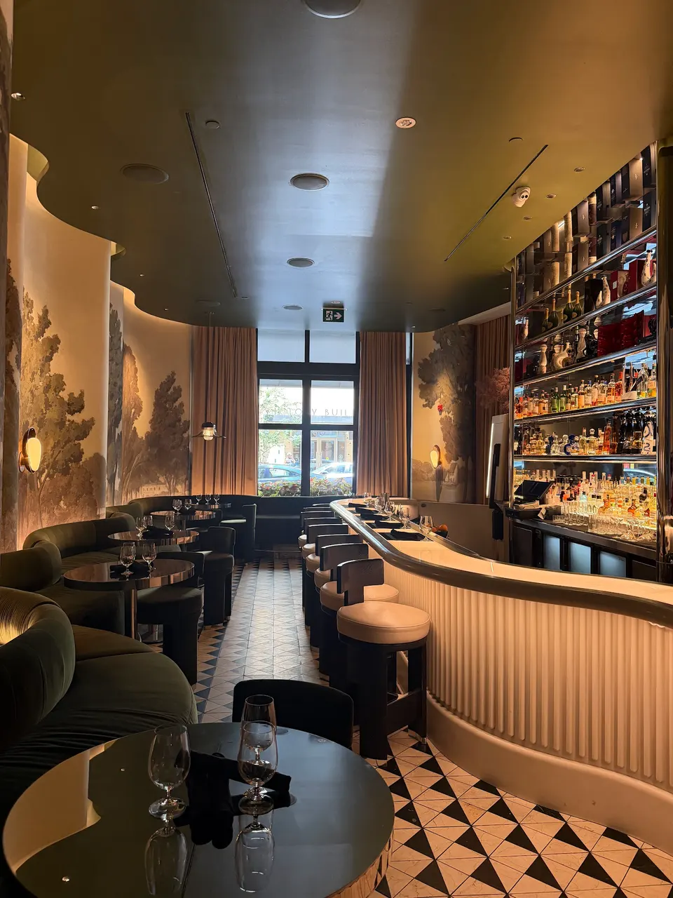 An elegant, dimly lit bar and dining area with curved green velvet booths, a long fluted bar counter with stools, and a scenic wall mural under a dark green ceiling.