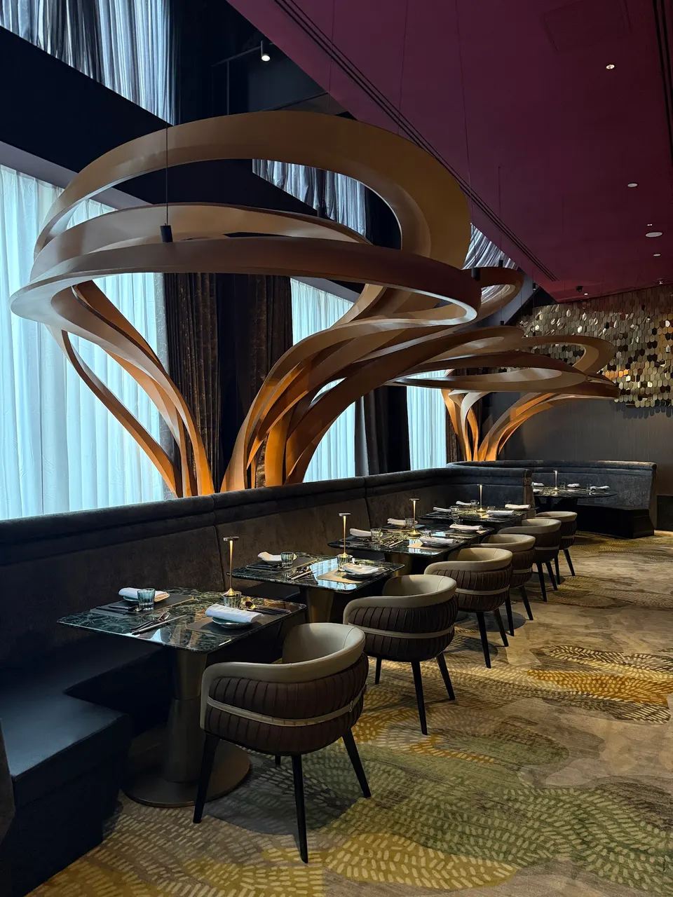 An upscale dining room featuring dark green marble tables, plush curved chairs, and a long dark banquette under large, sculptural wooden tree-like installations.