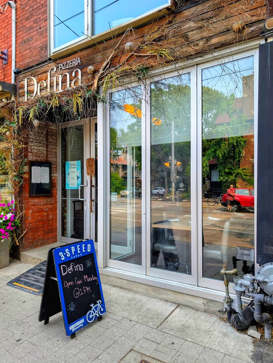 The exterior storefront of Pizzeria Defina featuring a rustic wooden sign, large glass folding doors, and a blue sandwich board sign on the sidewalk announcing holiday hours.