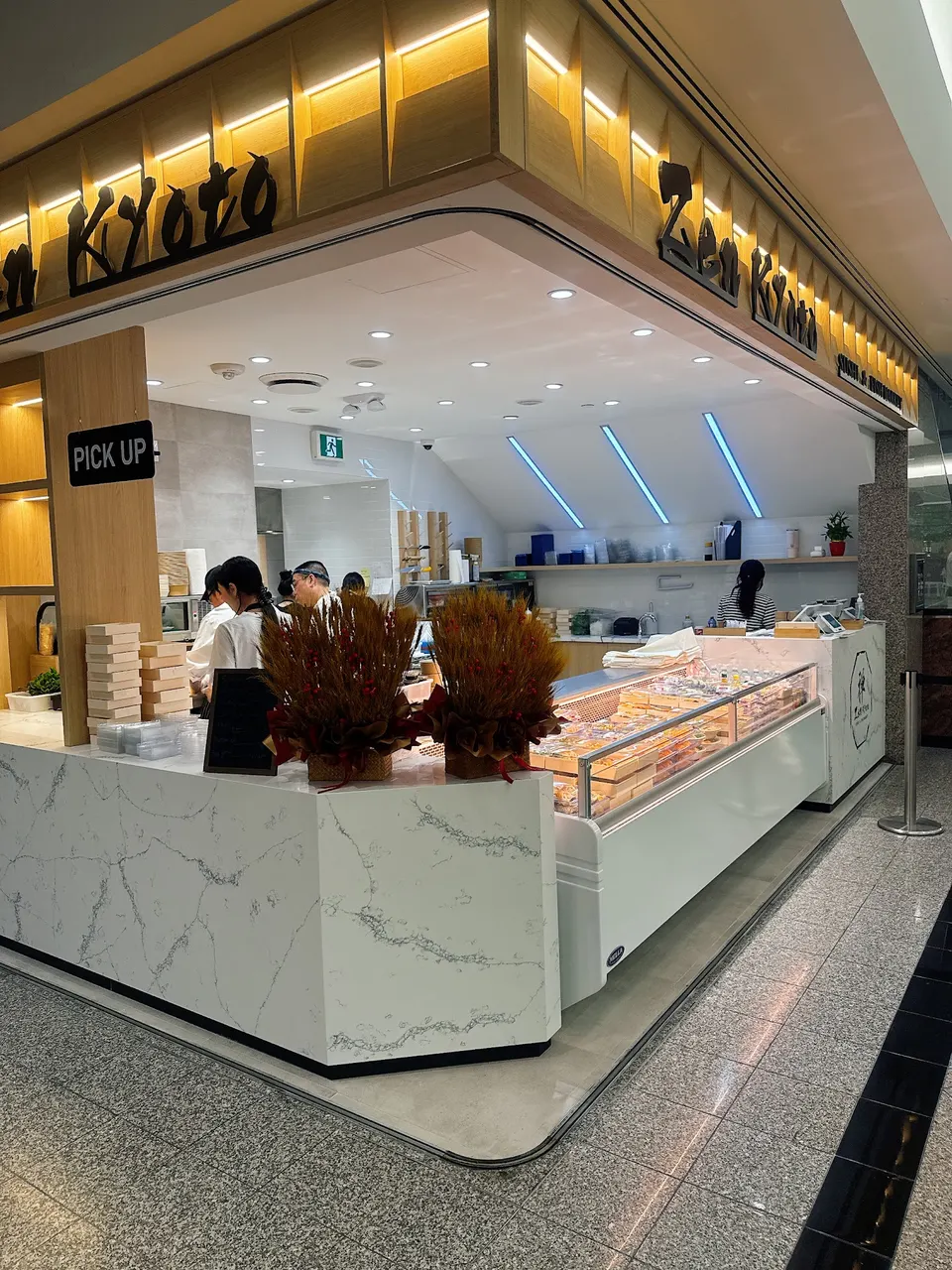 A modern Zen Kyoto food stall in a mall food court, featuring a white marble counter, a refrigerated display case filled with sushi boxes, and staff working behind the counter under bright recessed lighting.