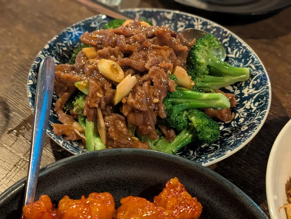 A plate of beef and broccoli stir-fry with garlic slices in a savory brown sauce, served on a blue and white patterned ceramic plate next to another dish.