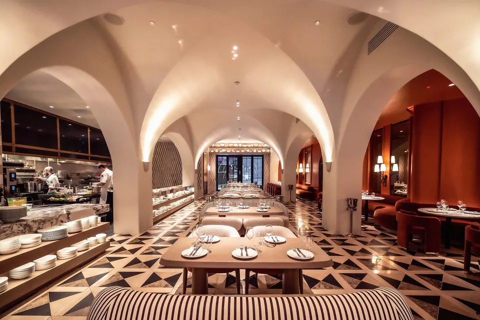 An elegant restaurant interior featuring white vaulted archways, patterned tile flooring, modern dining tables, and a glimpse of an open kitchen on the left.