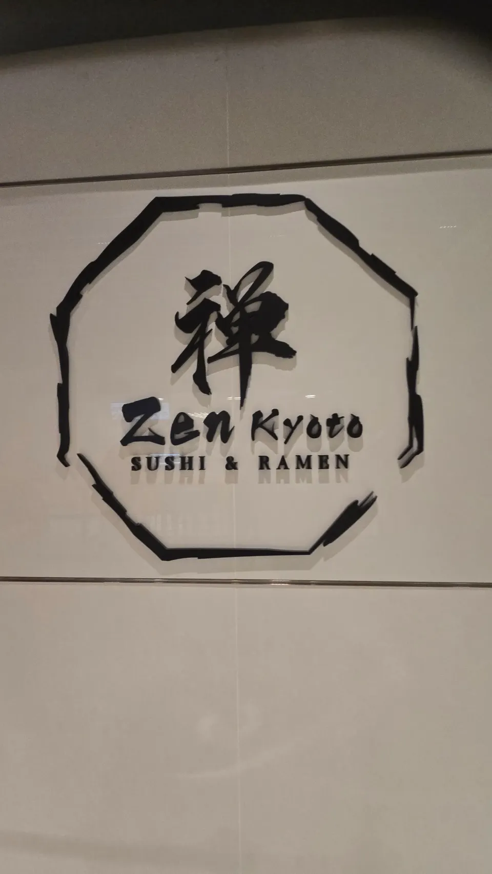 A white wall sign featuring a black octagonal brushstroke border enclosing a large Japanese kanji character for Zen, with the words 'Zen Kyoto' and 'SUSHI & RAMEN' written below.